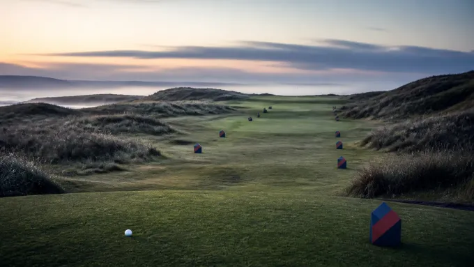 Original editorial illustration of one golf ball and navy-red route markers on an empty Irish links course at dawn.